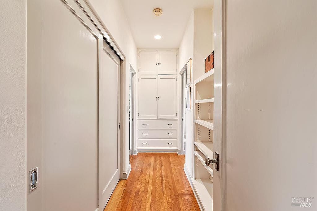 This interior hallway features warm-toned wood flooring and white walls, creating a bright and clean aesthetic. At the end of the hall, a built-in storage unit provides ample cabinet and drawer space, while an open shelving unit lines the right wall. The perspective is a straight-on view from a doorway, highlighting the functional layout and storage potential of the space.