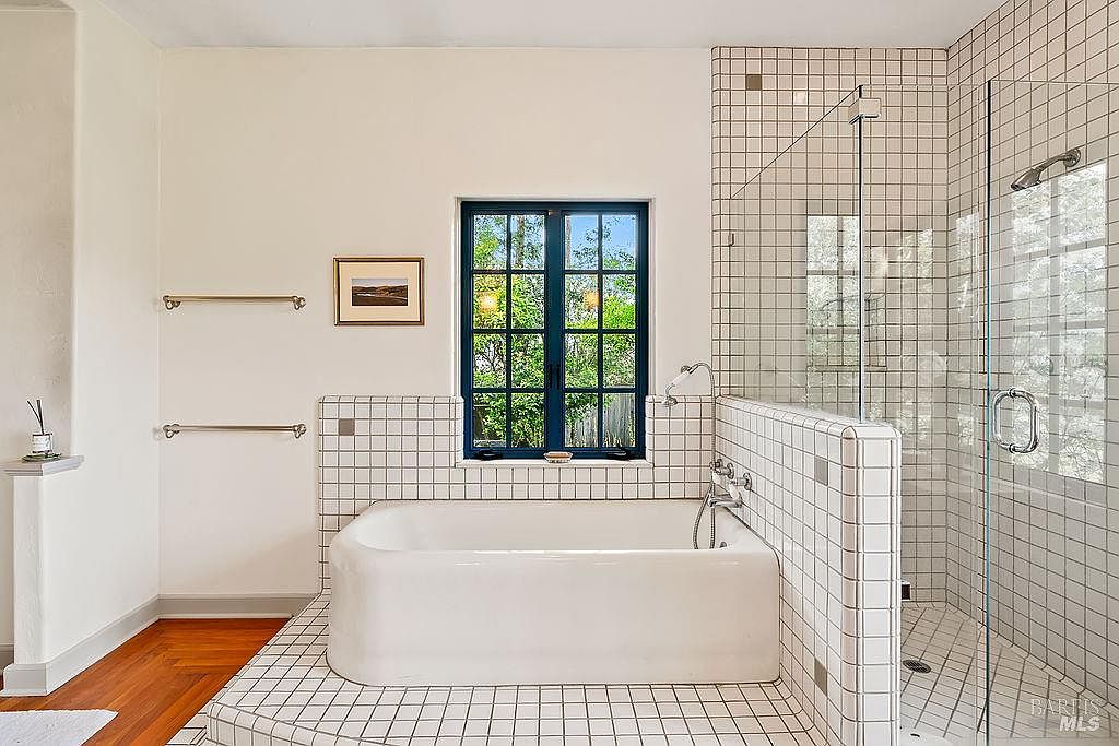 This bright bathroom features a classic white soaking tub set into a tiled platform, complemented by a walk-in shower with a glass enclosure and matching white square tiles. A dark-framed window with multiple panes provides natural light, while two wall-mounted towel bars add functional detail to the space. The room is finished with warm wood flooring, creating a clean and timeless aesthetic.