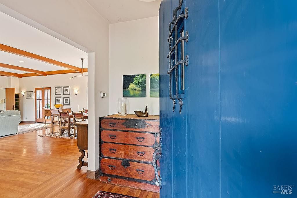 This entryway view captures a striking blue door with a decorative iron grille, opening into a warm, inviting home. Beyond the threshold, a rustic wooden chest sits against a wall, topped with a small bronze sculpture and framed artwork, leading the eye into a spacious living and dining area with exposed wood ceiling beams and polished hardwood floors. The perspective is from the doorway looking inward, highlighting the transition between the bold entry and the open-concept living space.