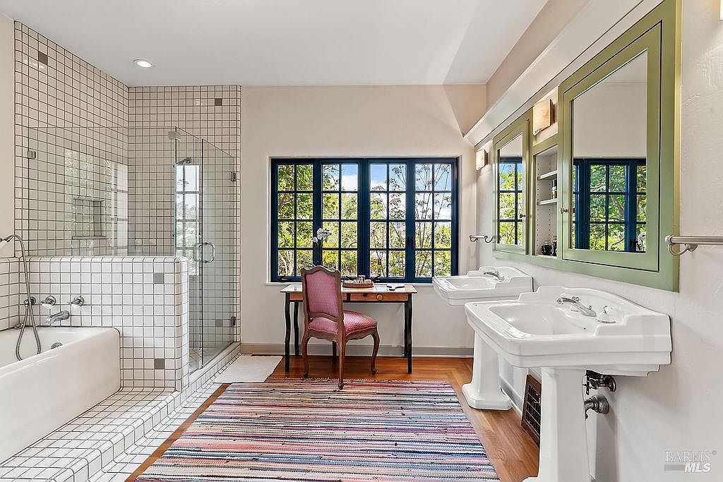 This spacious bathroom features a unique layout with a walk-in shower enclosed by glass, a separate bathtub, and a vanity area with two pedestal sinks. A desk and chair are positioned beneath a large window, adding a functional workspace to the room. The space is finished with white square wall tiles, wood flooring, and a colorful striped rug, creating a bright and eclectic atmosphere.