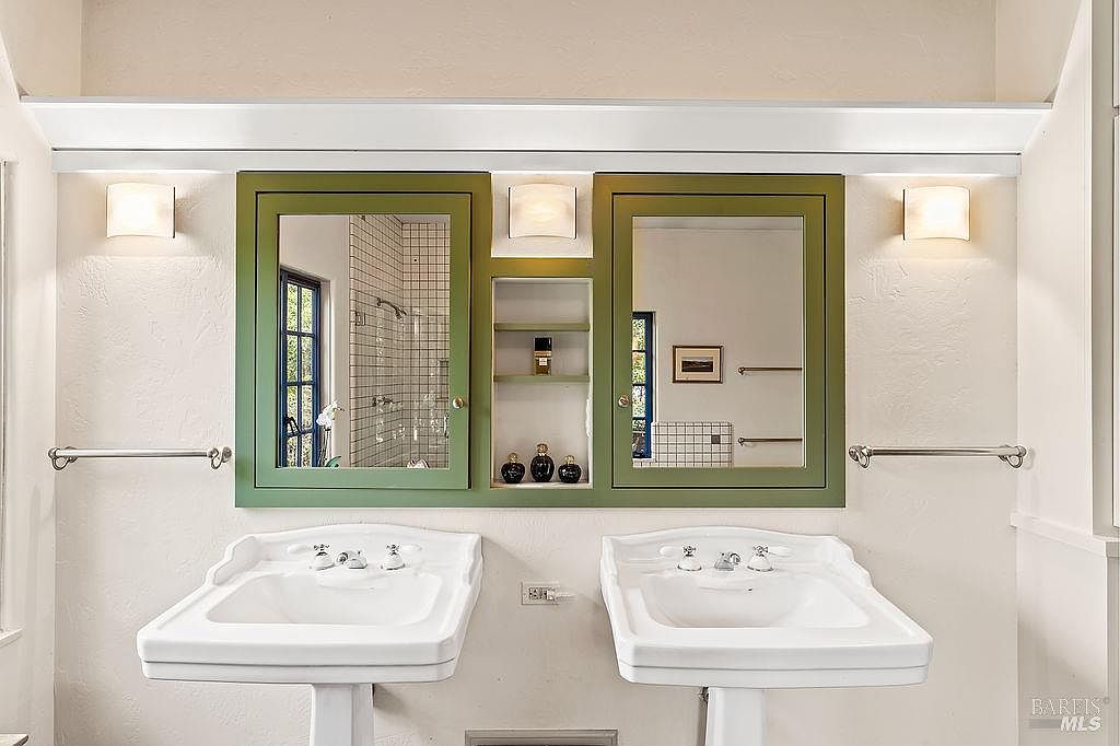 This bathroom features a double-sink vanity setup with two pedestal sinks positioned beneath a custom green-framed mirror and shelving unit. The central storage area includes open shelving for toiletries, flanked by two rectangular mirrors and wall-mounted light fixtures. The space is finished with neutral walls and chrome hardware, creating a classic and functional aesthetic.