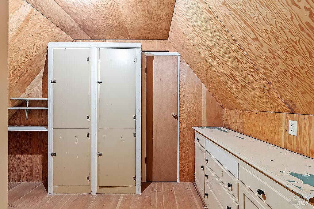 This attic space features sloped walls finished in light wood paneling, creating a cozy and rustic atmosphere. The room includes a built-in storage unit with two tall cabinet doors, a separate single door, and a long, low-profile dresser with multiple drawers along the right wall. The space is illuminated by a single wall-mounted electrical outlet and appears to be a functional storage or utility area.