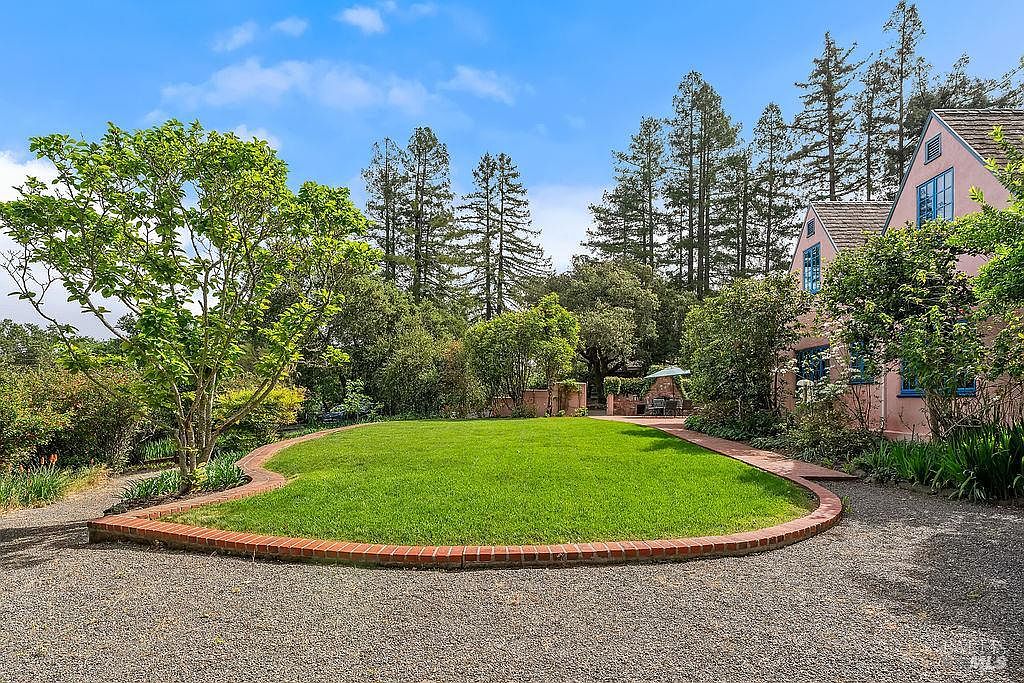 This image captures a serene backyard featuring a manicured, oval-shaped lawn bordered by a classic red brick edge. To the right, a soft pink house with blue-trimmed windows stands amidst lush greenery, while a gravel path leads toward a distant outdoor seating area. The scene is framed by tall, mature evergreen trees under a bright blue sky, creating a peaceful and private atmosphere.