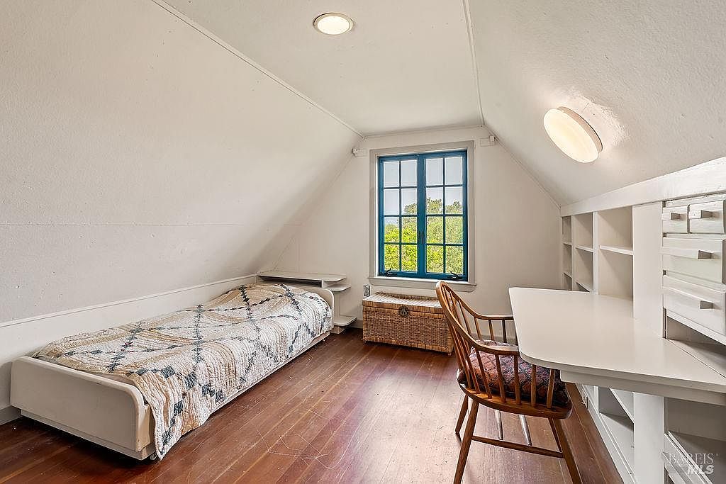 This cozy attic bedroom features sloped ceilings and warm wood flooring, creating a charming and intimate atmosphere. The room is furnished with a single bed, a built-in desk with shelving, and a wicker storage chest placed beneath a window that offers a view of the outdoors. The space is illuminated by a recessed ceiling light and a wall-mounted fixture, highlighting the clean, light-toned walls.
