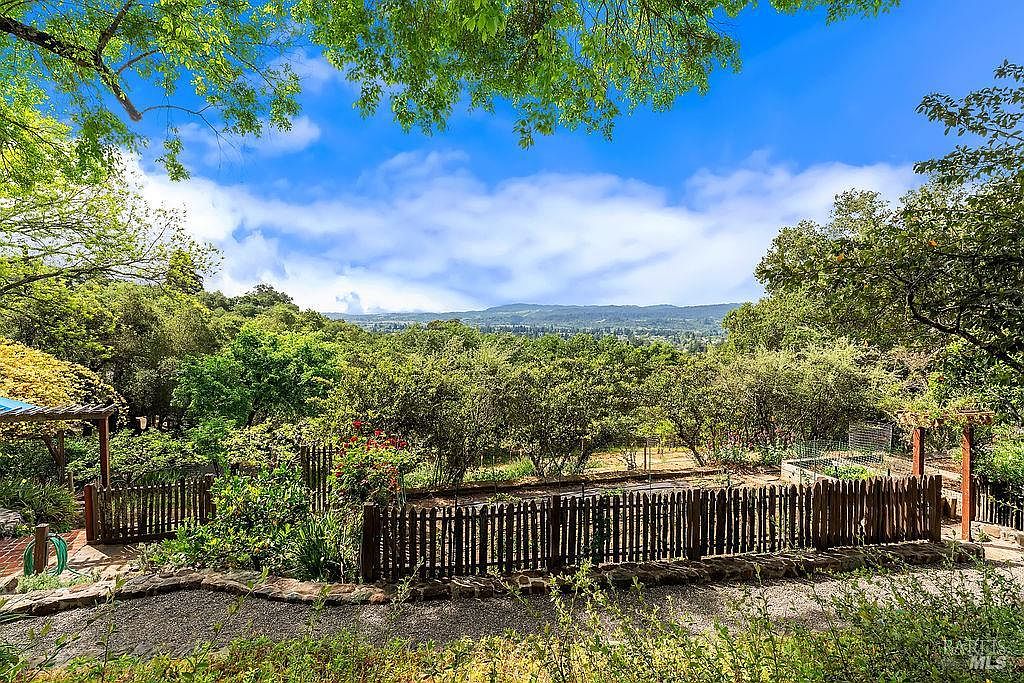 This scenic outdoor space features a fenced garden area set against a backdrop of lush, rolling hills and a vibrant blue sky. A gravel path leads toward the garden, which is framed by mature trees and a rustic wooden fence, creating a peaceful and private atmosphere. The perspective is captured from a slightly elevated position, emphasizing the expansive natural landscape and the well-maintained garden beds.