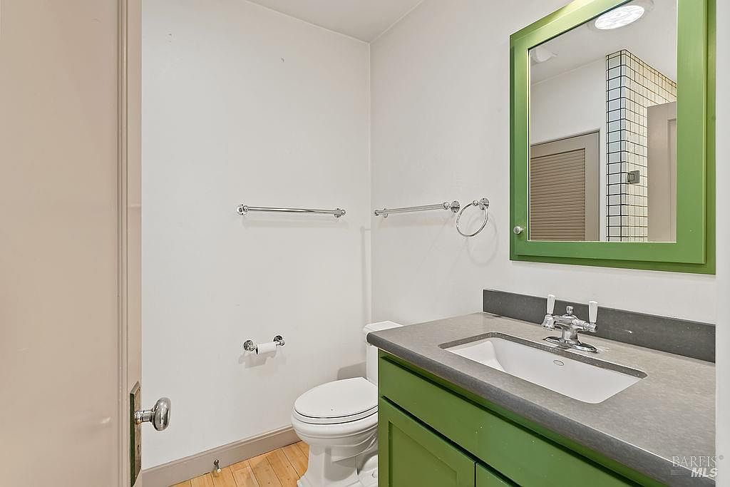This compact bathroom features a vanity with a green cabinet base and a gray countertop, complemented by a matching green-framed mirror. The space includes a white toilet, a wall-mounted towel bar, and a toilet paper holder, all set against plain white walls and light wood flooring. The perspective is a straight-on view from the doorway, highlighting the functional layout of this small powder room.