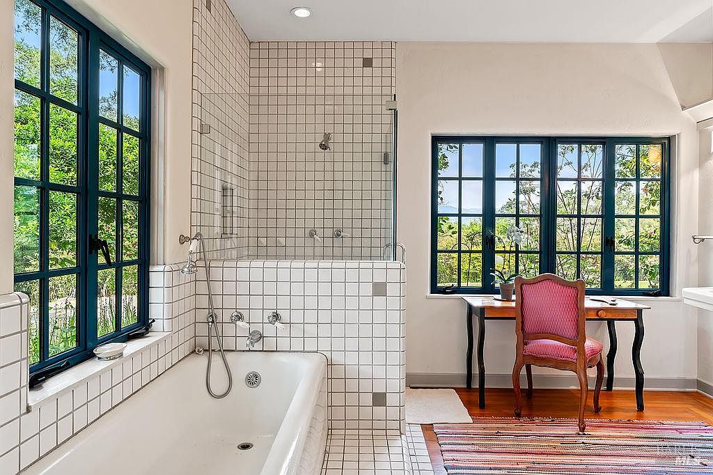 This bright and airy bathroom features a classic white-tiled bathtub and shower combination with a glass enclosure. A wooden desk with a pink upholstered chair is positioned near large, multi-pane windows that offer a view of the lush greenery outside. The space combines functional bathing facilities with a unique study area, creating a serene and versatile atmosphere.