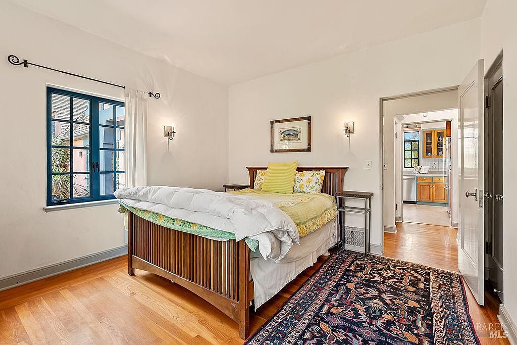 This bedroom features a wooden bed frame centered against a wall adorned with a framed picture and two wall-mounted sconces. A large, dark-framed window with blue trim allows natural light to enter, while a patterned area rug adds texture to the light wood flooring. The room opens into a bright kitchen area, creating an open and connected feel between the living and utility spaces.