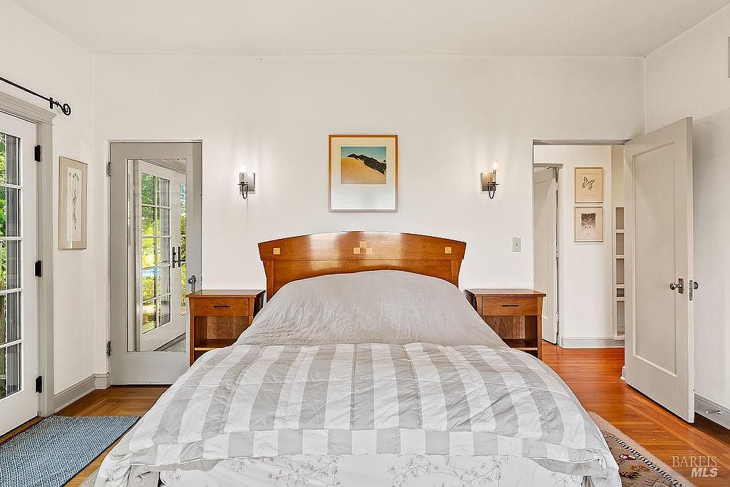This bedroom features a central bed with a warm-toned wooden headboard flanked by matching nightstands and wall-mounted sconces. The room is bright and airy, with white walls, hardwood flooring, and a glass-paned door that provides a view of the outdoors. A framed piece of art hangs above the bed, and an open doorway leads to an adjacent hallway, creating a sense of flow and openness.