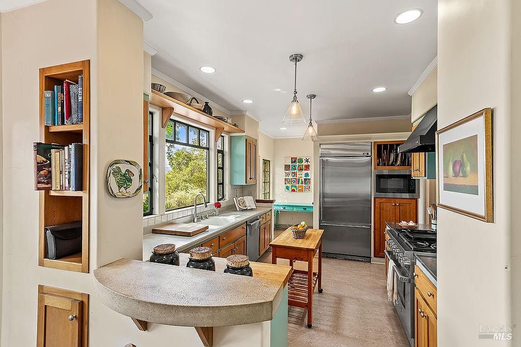 This bright and inviting kitchen features a functional layout with a central wooden island, stainless steel appliances, and a breakfast bar with a stone-topped counter. The space is accented by warm wood cabinetry, open shelving for books and decor, and a window above the sink that brings in natural light. The perspective is from the living area looking into the kitchen, highlighting the open-concept design and cozy, lived-in atmosphere.