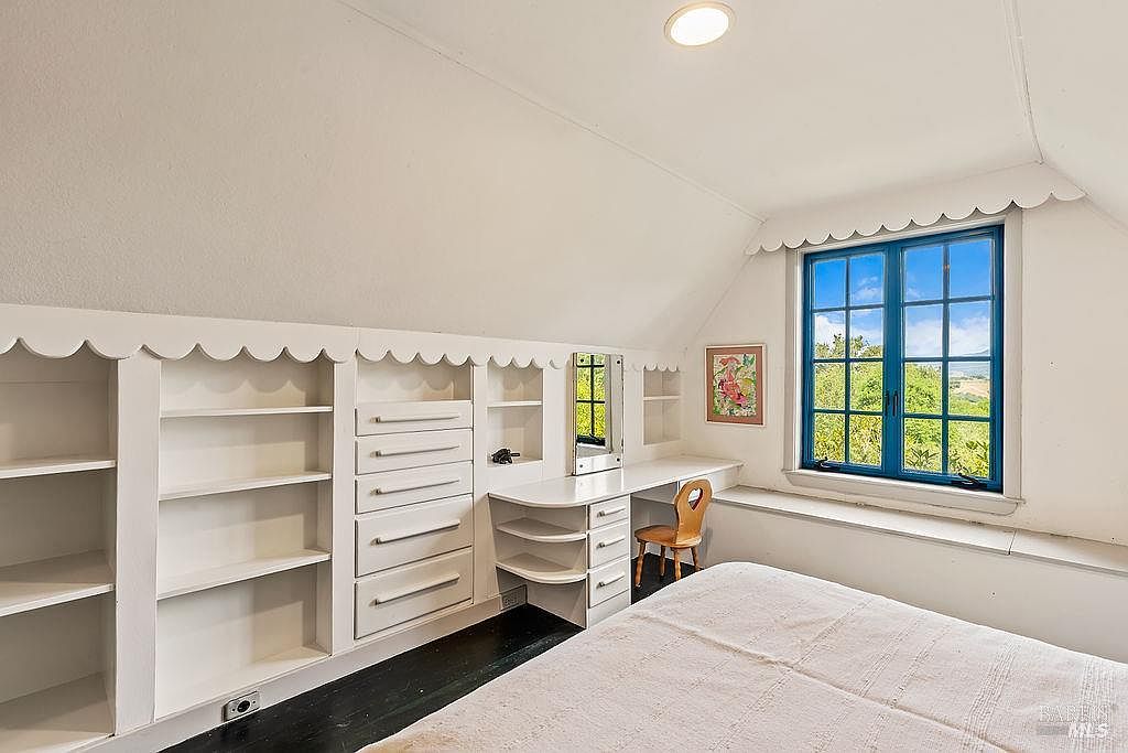 This cozy attic bedroom features a sloped ceiling with a charming scalloped trim detail along the built-in storage units. The room includes a desk area with a mirror, a set of drawers, and open shelving, all finished in white, contrasting with the dark wood flooring. A window with blue frames provides a view of the outdoors, and a simple wooden chair sits at the desk, creating a quaint and functional living space.