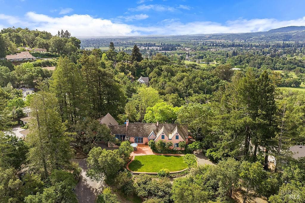This high-angle aerial view captures a charming, multi-gabled home nestled within a lush, wooded landscape. The property features a well-manicured lawn, a terracotta-toned patio area, and a classic architectural style that blends seamlessly with the surrounding mature trees and rolling hills in the distance. The perspective provides a comprehensive look at the home's roofline, layout, and its serene, private setting.