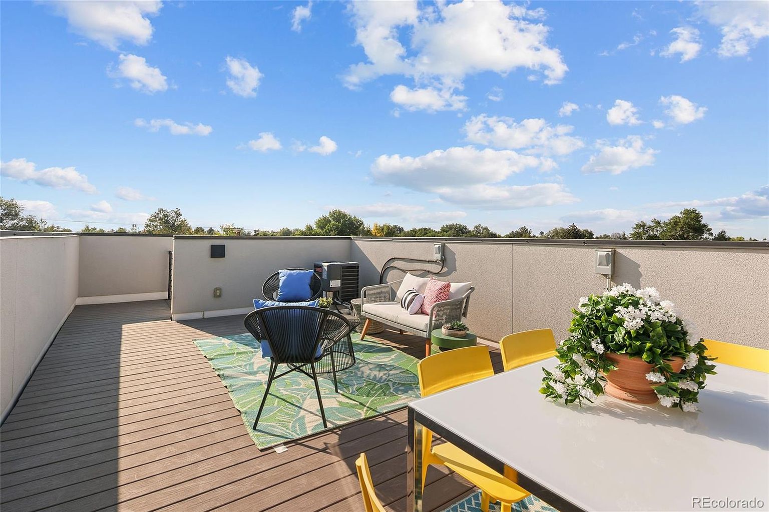 This image showcases a stylish rooftop patio or deck area, furnished with modern outdoor furniture including a dining table with yellow chairs and a lounge area with comfortable seating. The deck is made of wood planks, and a green patterned rug adds a pop of color. The view extends to a blue sky with scattered clouds, creating an inviting and relaxing outdoor space.