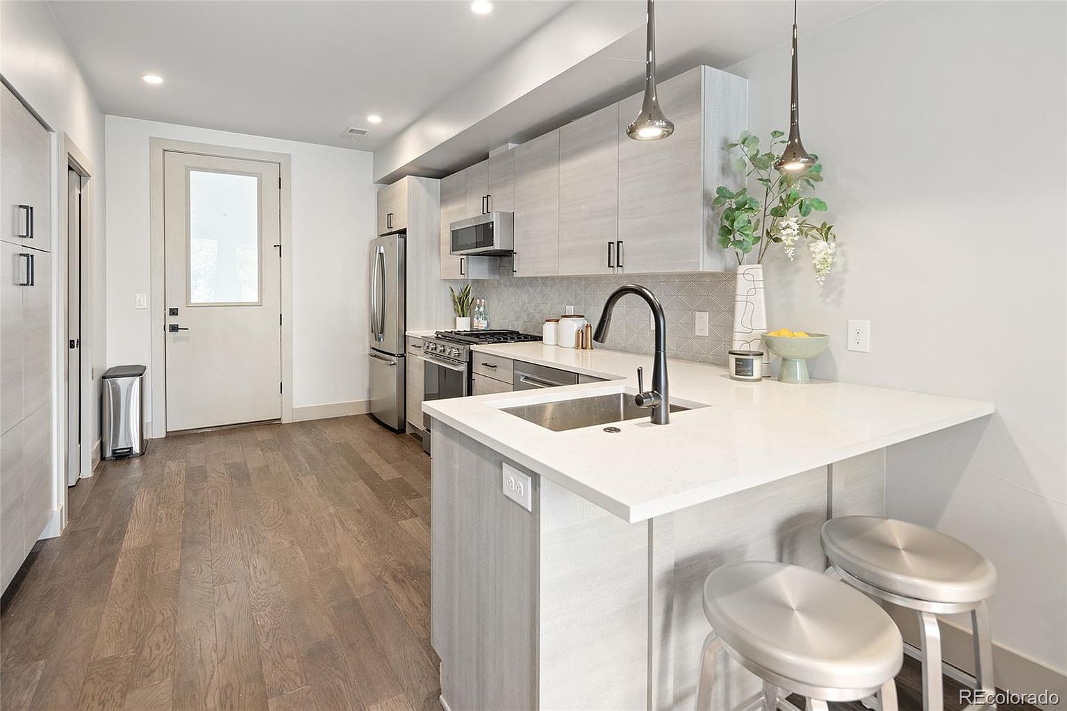 This is a well-lit kitchen with modern design elements. It features light wood-grain cabinets, stainless steel appliances, and a white countertop island with seating. The kitchen also has pendant lighting and a door leading to another area, creating a clean and inviting space.