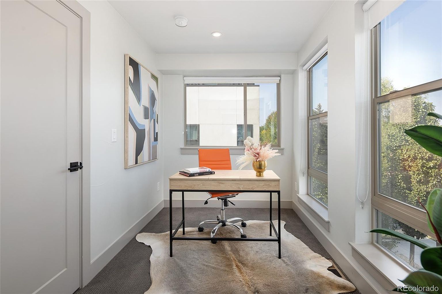 This is an interior shot of a modern office space. The room features a light wood desk with a black metal frame, an orange office chair, and a cowhide rug. Natural light floods the room through large windows, and an abstract painting hangs on the wall, creating a bright and inviting workspace.