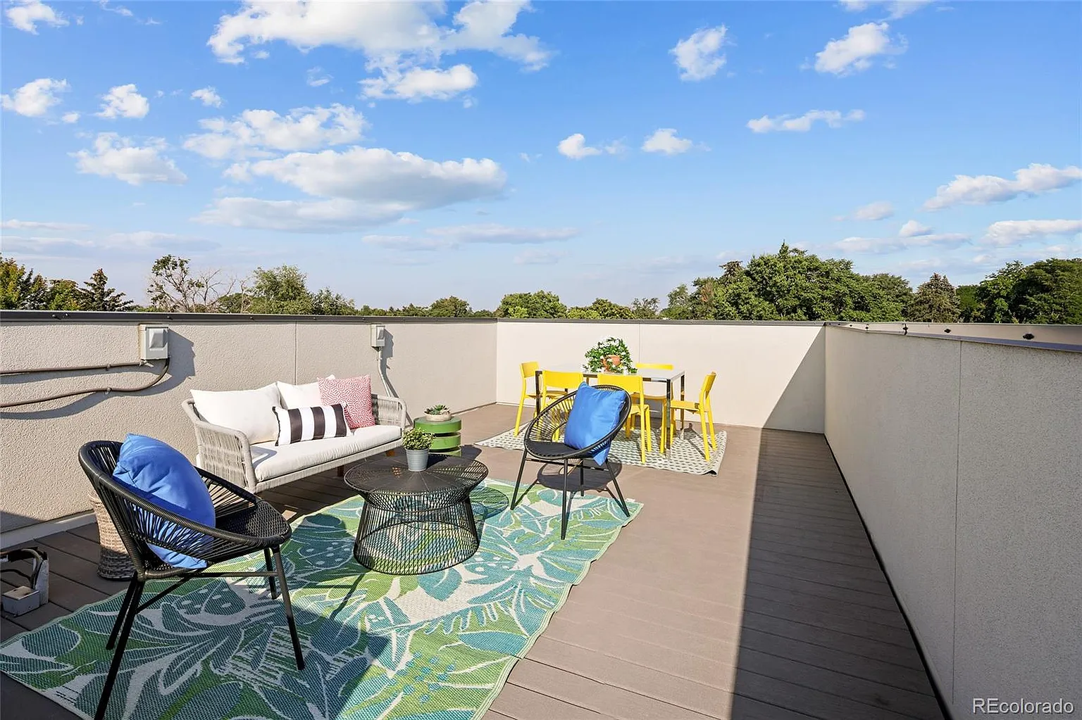 This image showcases a well-appointed rooftop patio featuring stylish outdoor furniture and decor. The patio includes a seating area with a sofa and chairs, a dining table set, and a colorful rug that adds visual appeal. With its inviting arrangement and neutral backdrop, the space is ideal for both relaxation and entertaining.