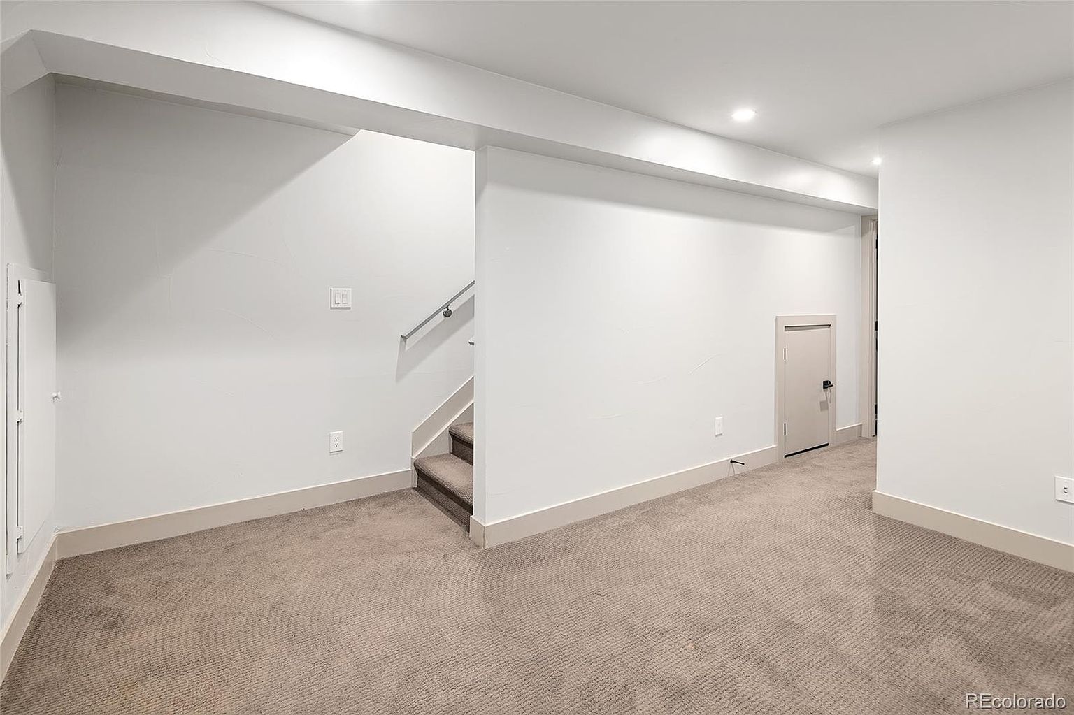 The image showcases a clean and well-lit basement area with neutral-toned carpet and white walls. A staircase is visible, suggesting access to another level, and a small door is present, possibly leading to a storage area or utility room. The space appears versatile and ready for customization, offering potential as a living area, home office, or recreational space.