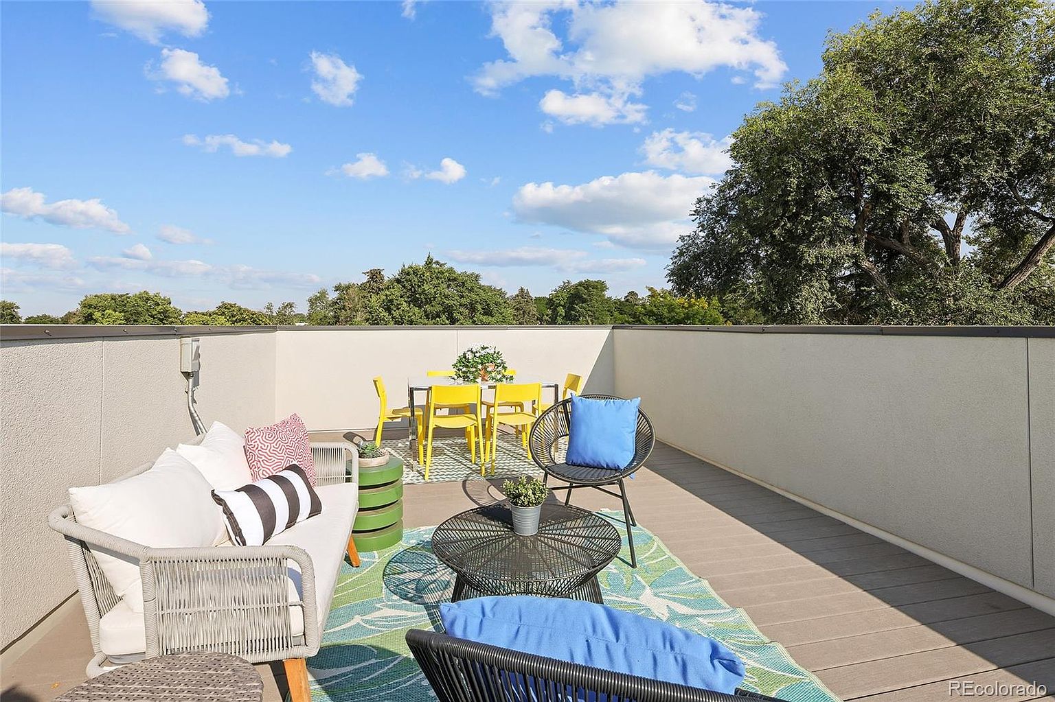 This image showcases a stylish outdoor patio or balcony area, furnished with modern seating, a dining table with yellow chairs, and a decorative rug. The space is enclosed by a white wall, offering privacy, while the open sky and surrounding trees provide a pleasant backdrop. The overall impression is one of a comfortable and inviting outdoor living space.