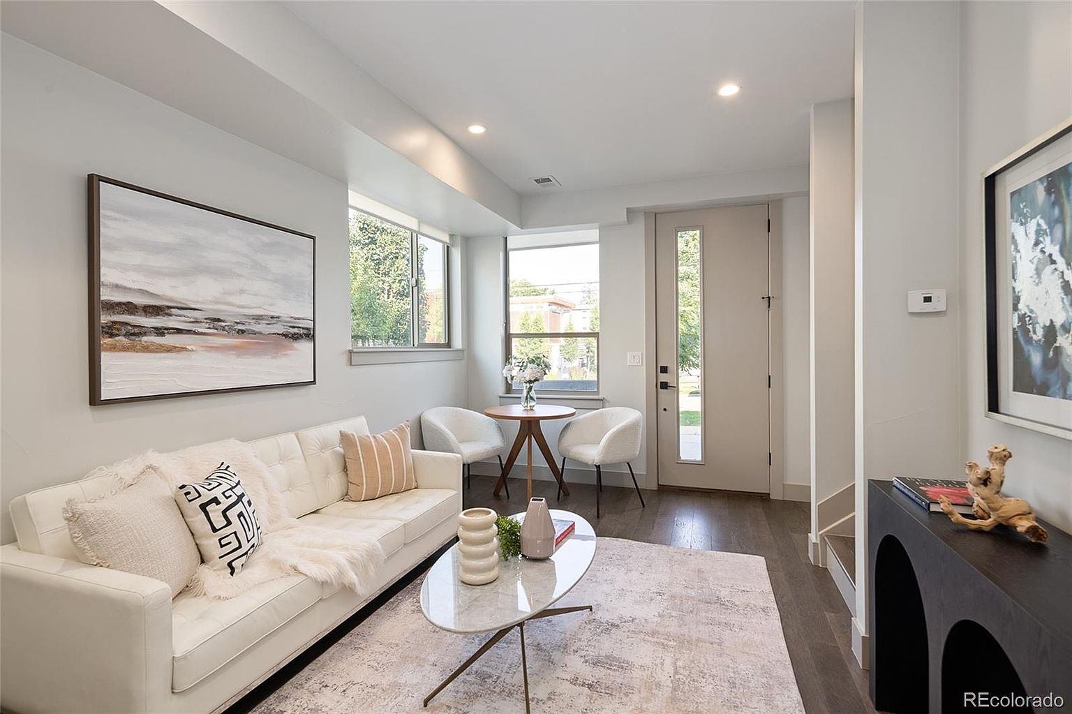 This is an interior shot of a living room featuring a white sofa with decorative pillows, a glass-topped coffee table with vases, and two white chairs around a small round table. The room has a modern aesthetic with neutral colors, hardwood floors, and natural light coming through the windows, creating a bright and inviting space.