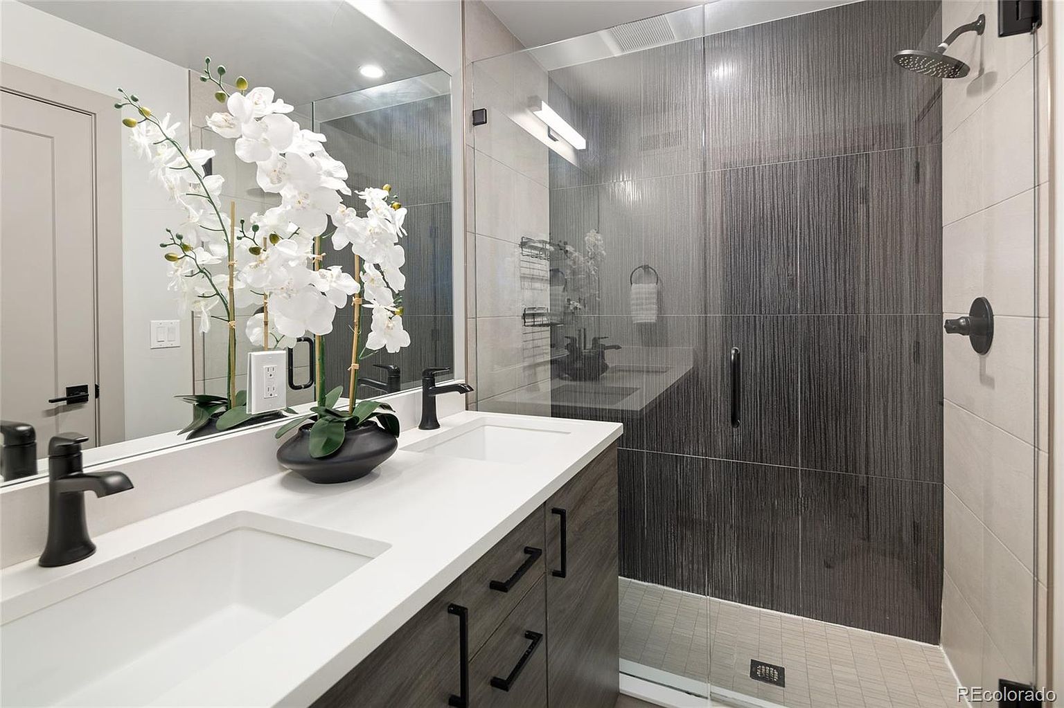 This is a well-lit primary bathroom featuring a double vanity with white countertops and dark wood cabinetry. Black fixtures and a large mirror enhance the modern aesthetic. A glass-enclosed shower with contrasting light and dark tile adds a luxurious touch, creating a spa-like atmosphere.