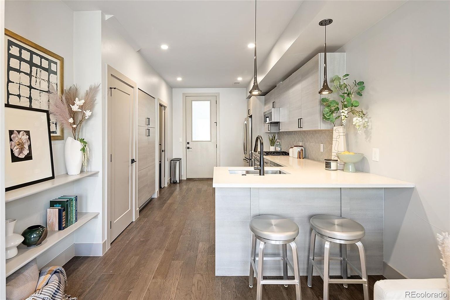 This is a bright and modern kitchen featuring light wood-tone cabinetry, stainless steel appliances, and a white countertop island with seating. Pendant lights hang above the island, and the open layout connects seamlessly to the hallway. The space is clean, well-lit, and inviting, perfect for cooking and entertaining.