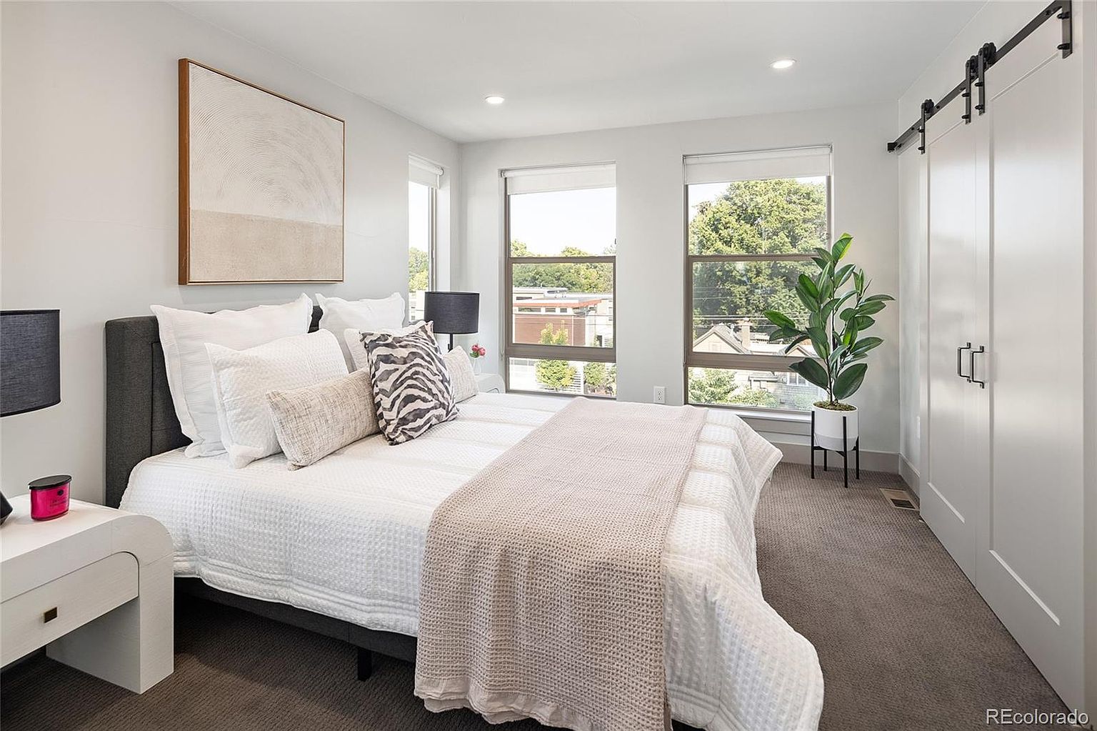 This is a bright and airy primary bedroom featuring a large bed with multiple pillows and a throw blanket. The room has two large windows that let in natural light, complemented by a modern art piece on the wall. A sliding barn door adds a touch of rustic charm to the contemporary design.