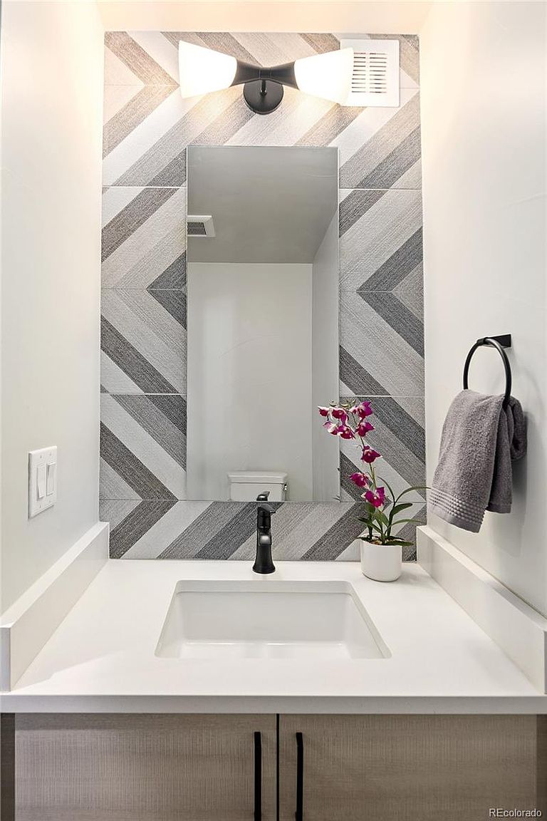 This is a well-lit guest bathroom featuring a modern design. The vanity has a white countertop, a rectangular sink, and light wood-tone cabinets with black hardware. A decorative tile backsplash with a chevron pattern adds visual interest, and a black faucet and towel ring provide a sleek contrast.
