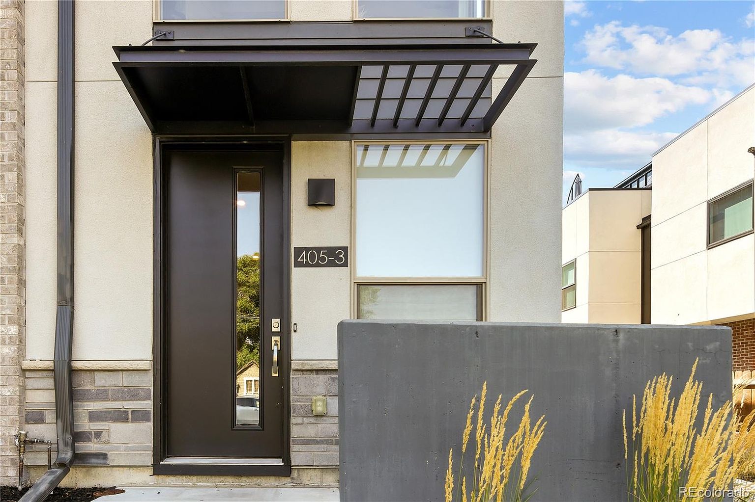This image showcases the entryway of a modern home, featuring a dark-colored front door with a vertical window pane and a sleek, geometric awning. The house number '405-3' is prominently displayed next to the door, and a contemporary concrete planter with ornamental grasses adds a touch of landscaping. The overall impression is clean, stylish, and inviting.