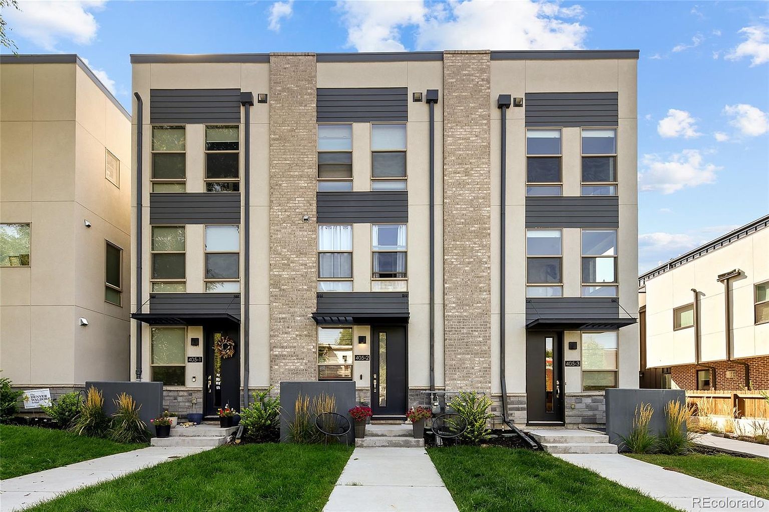 This is a front view of three modern townhouses. The townhouses feature a combination of beige siding and brick accents, with dark gray trim and awnings. Each unit has a well-maintained lawn and a concrete walkway leading to the front door, creating a welcoming and contemporary curb appeal.