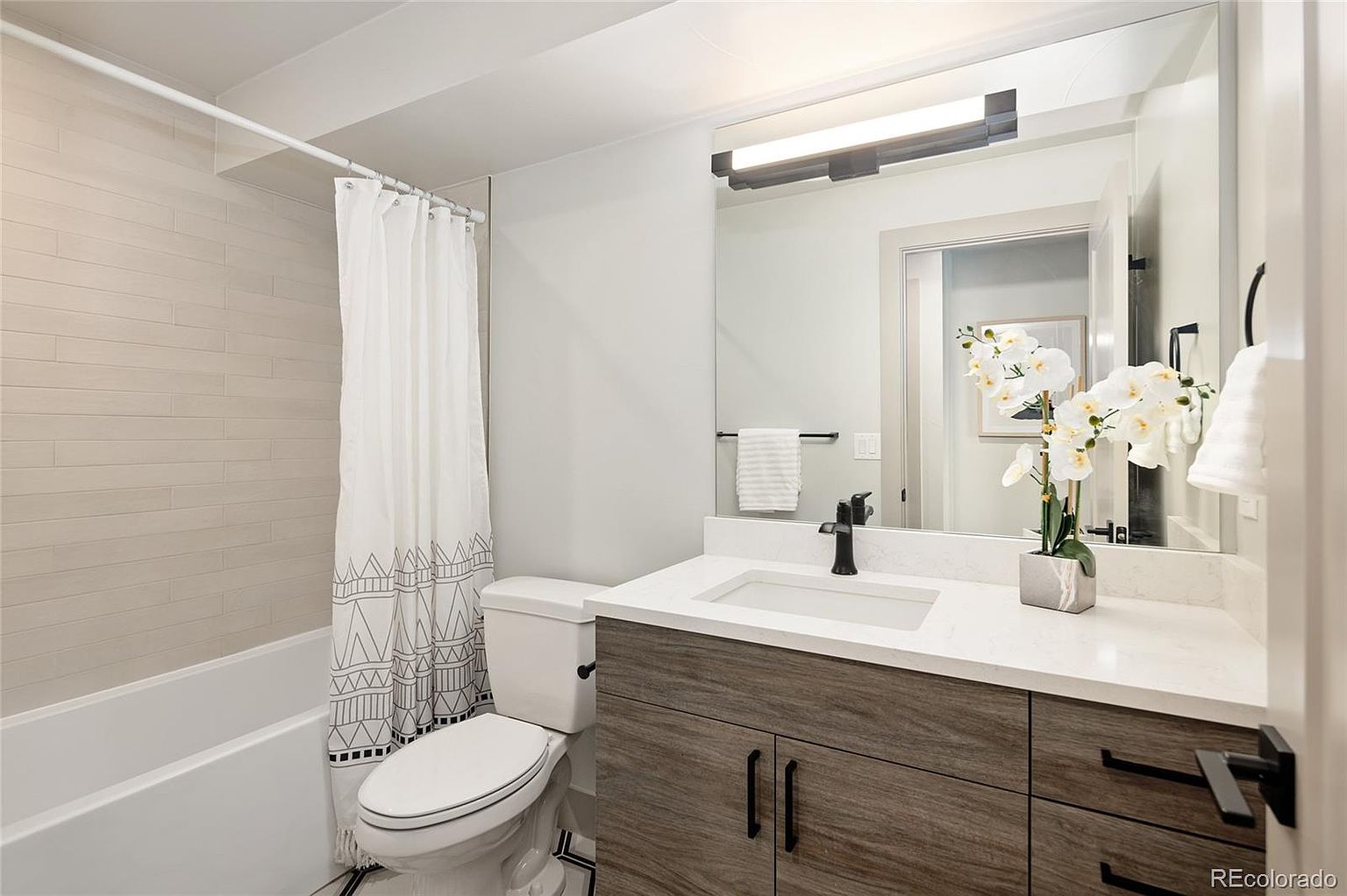 This is a well-lit bathroom featuring a modern design. The bathroom includes a toilet, a vanity with a white countertop and wood-grain cabinets, a black faucet, and a mirror with a light fixture above. A bathtub with a shower curtain is also visible, and the overall impression is clean and stylish.