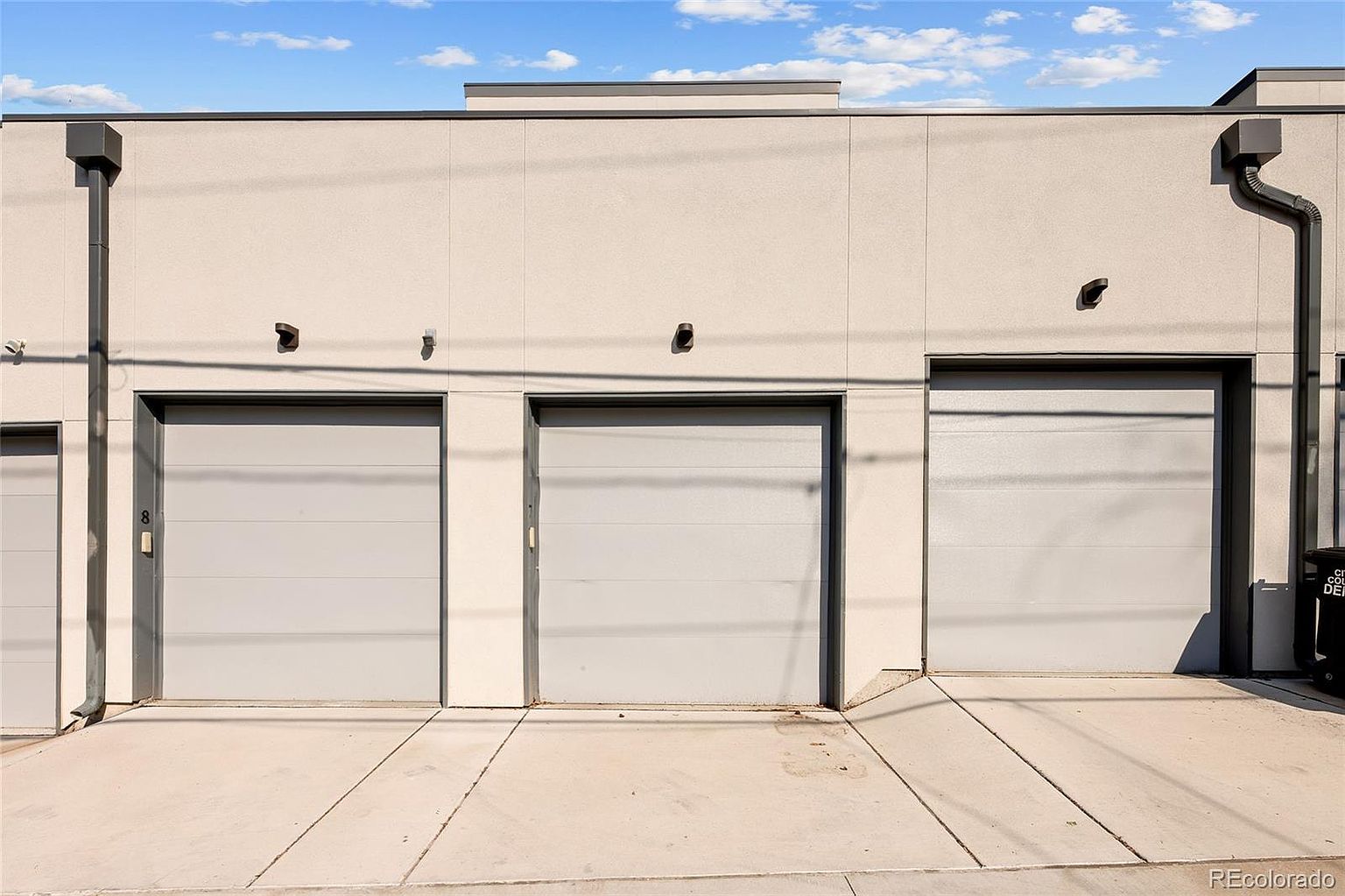 The image showcases the exterior of a building with multiple garage doors, all painted in a neutral gray tone. The building's facade is a light beige, and the overall design is modern and minimalist. The concrete driveway in front of the garages is clean and well-maintained, suggesting a functional and contemporary space.