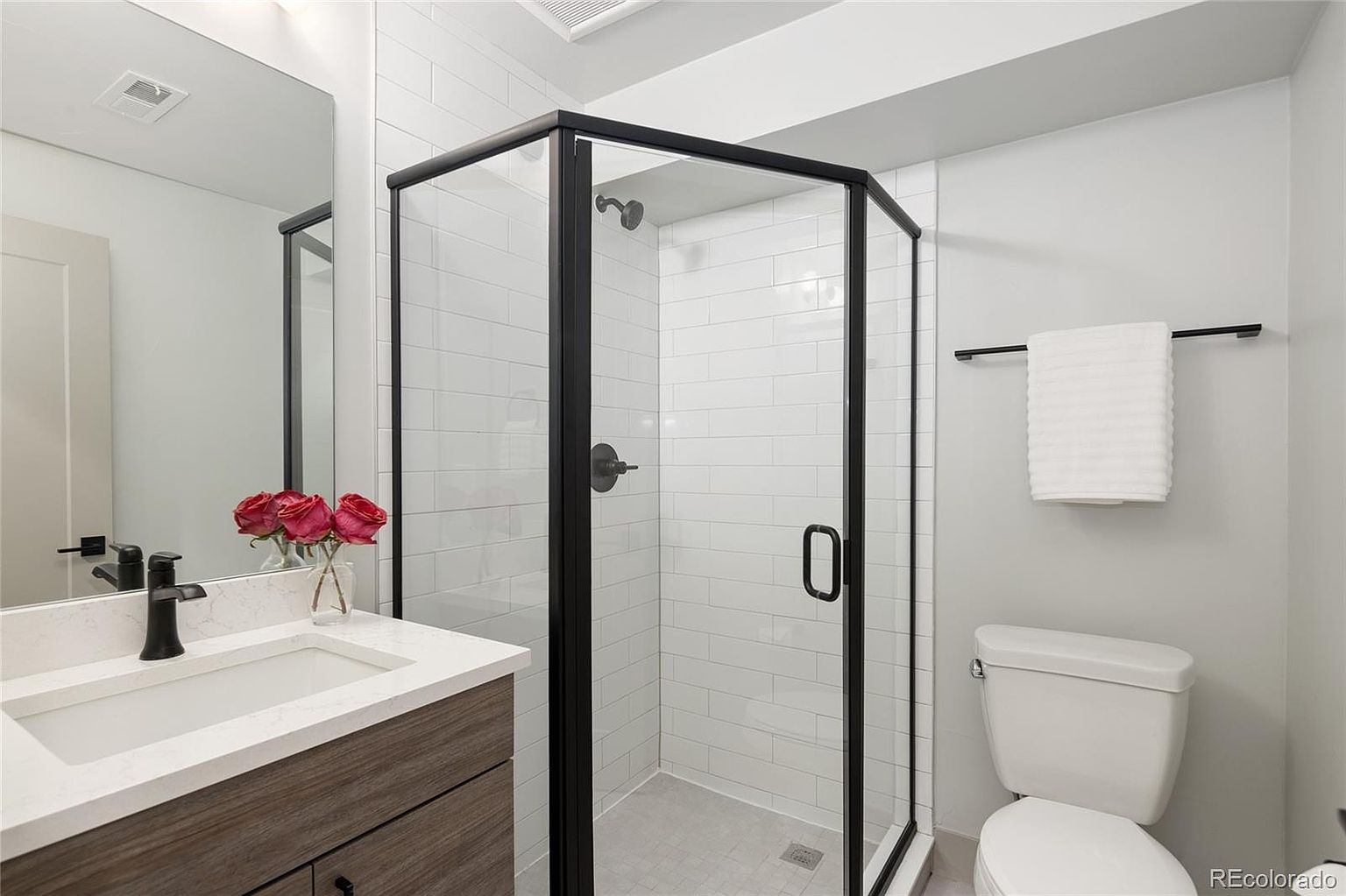 This is a well-lit bathroom featuring a modern design with a black-framed glass shower enclosure and white subway tile. The vanity has a light countertop and wood-tone cabinets, complemented by black hardware. A white toilet and a towel bar with a neatly folded white towel complete the space, creating a clean and inviting atmosphere.