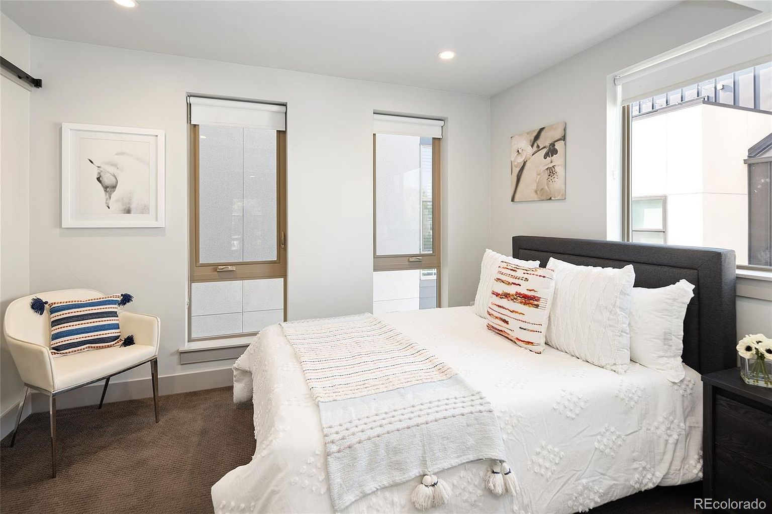 This is a bright and airy primary bedroom featuring a bed with a gray headboard, white bedding, and decorative pillows. Two windows with blinds provide natural light, and artwork adorns the walls. A chair with a decorative pillow sits near one of the windows, and a dark wood nightstand is next to the bed.
