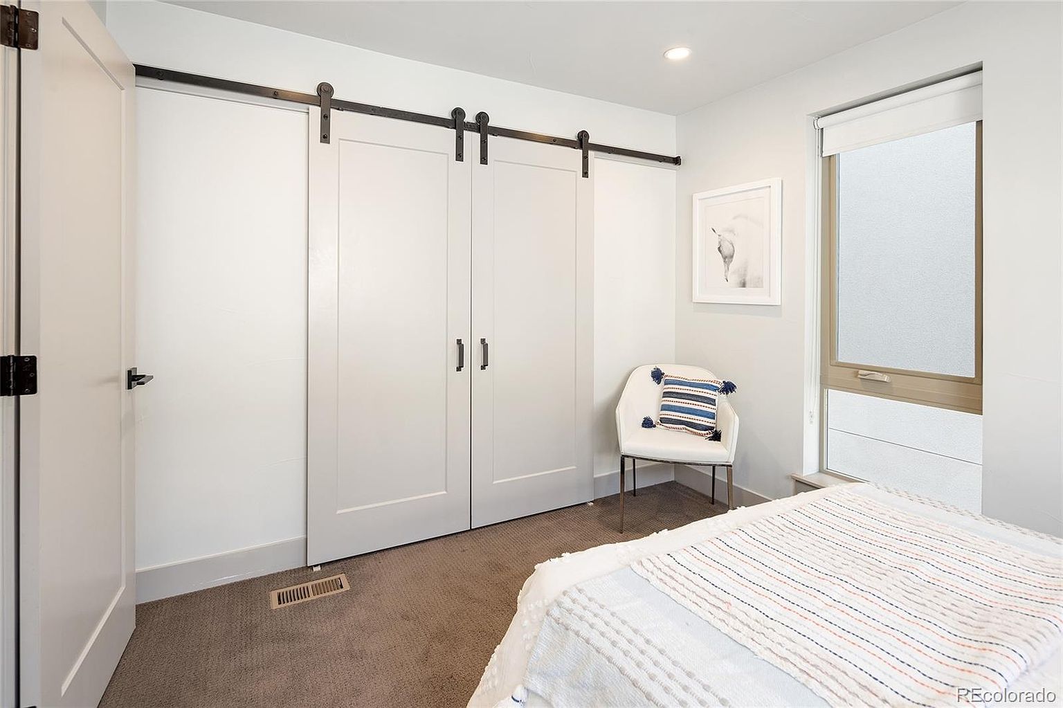 This is a bright and airy bedroom featuring a sliding barn door closet, a modern chair with a striped pillow, and a window with a shade. The walls are painted in a neutral tone, and the flooring appears to be carpeted. The room has a clean and minimalist aesthetic, creating a calming and inviting space.