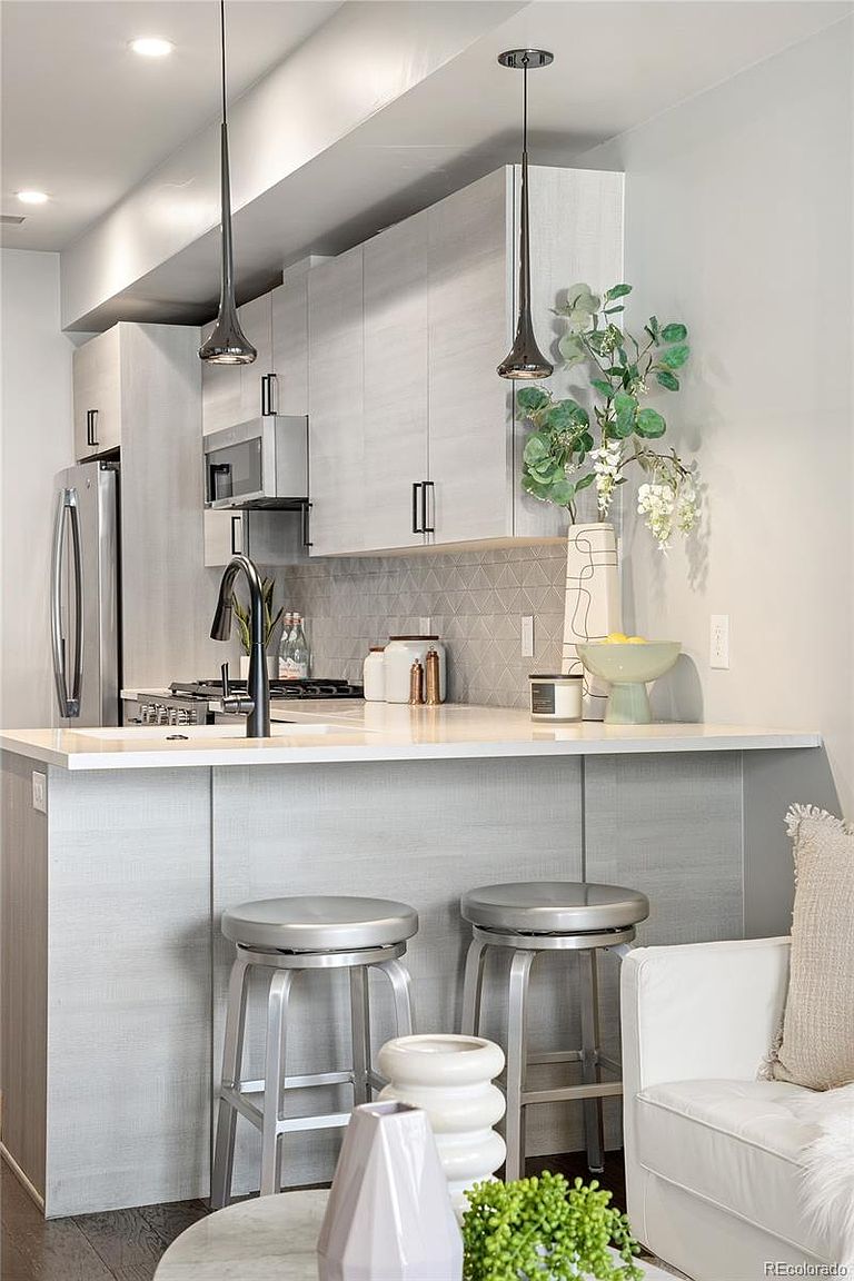 This is a modern kitchen with light gray cabinetry and stainless steel appliances. A white countertop island features two silver bar stools, and pendant lights hang above. The backsplash is a geometric pattern, and the overall impression is clean and contemporary.
