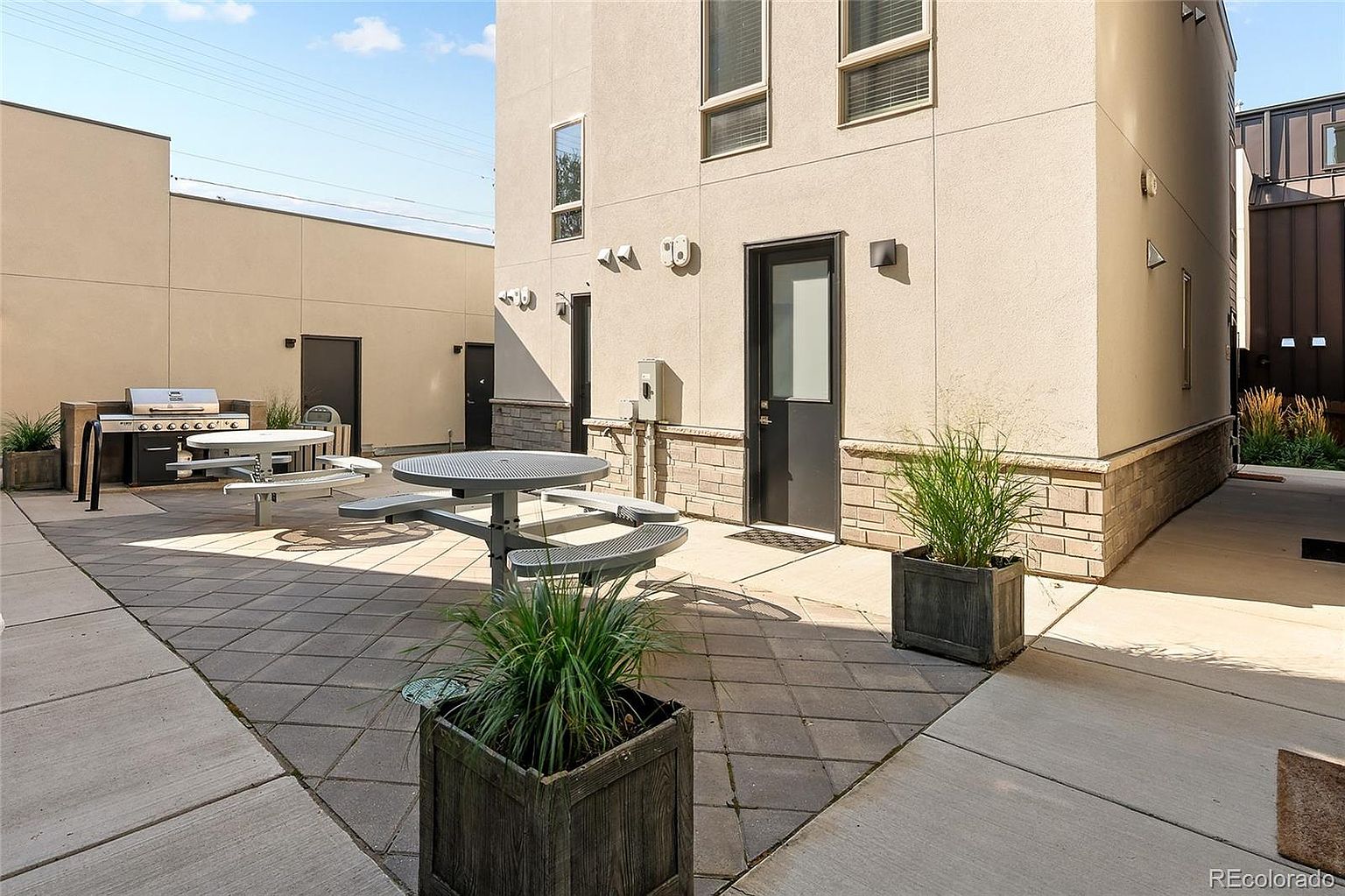 This image showcases a community outdoor amenity area, featuring picnic tables, a grill, and well-maintained landscaping. The area is paved with stone tiles and surrounded by modern buildings with neutral tones. The scene suggests a comfortable and inviting space for residents to gather and enjoy outdoor activities.