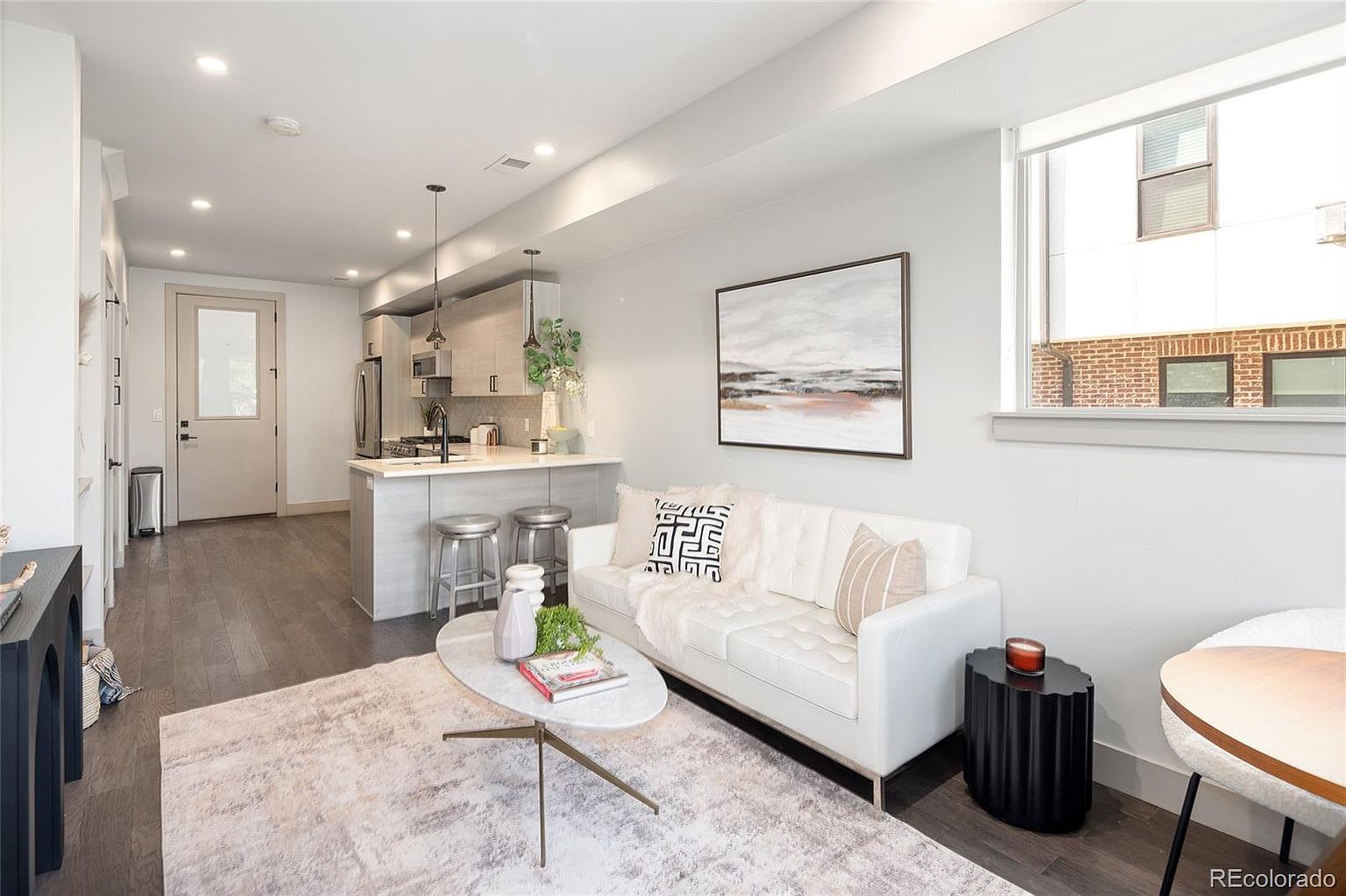 This is an interior shot of a modern living room and kitchen area. The living room features a white sofa, a light-colored rug, and a unique coffee table, complemented by a piece of artwork on the wall. The kitchen area includes a breakfast bar with seating, stainless steel appliances, and light wood cabinetry, creating a cohesive and stylish living space.
