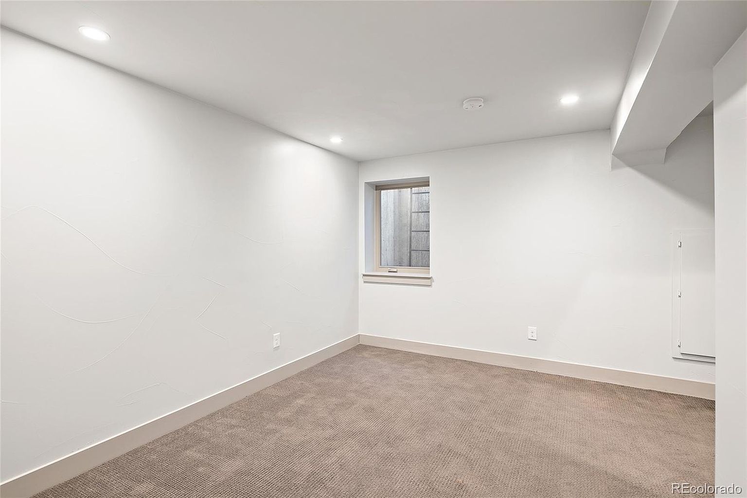 The image showcases a clean, finished basement space with neutral-toned carpet and white walls. A small window provides natural light, and recessed lighting illuminates the room. The space appears versatile and ready for various uses, such as a recreation room or home office.