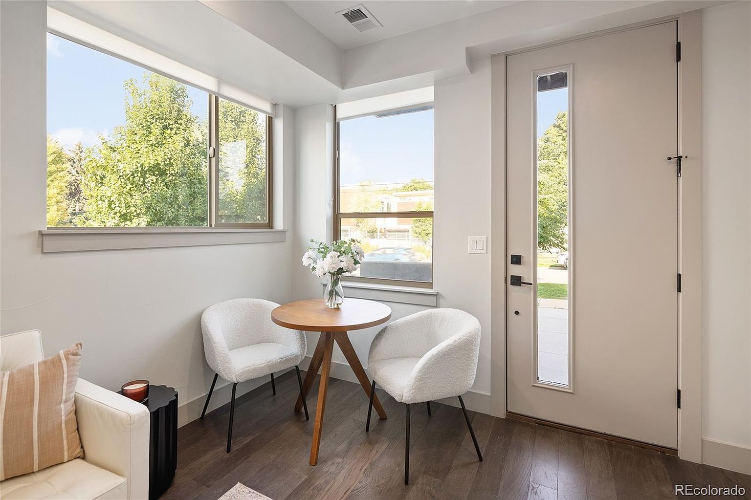 This interior shot showcases a cozy living area with a modern aesthetic. Two white chairs surround a small wooden table adorned with flowers, positioned near a window that offers natural light and a view of the outdoors. The room features hardwood floors and a neutral color palette, creating a serene and inviting atmosphere.