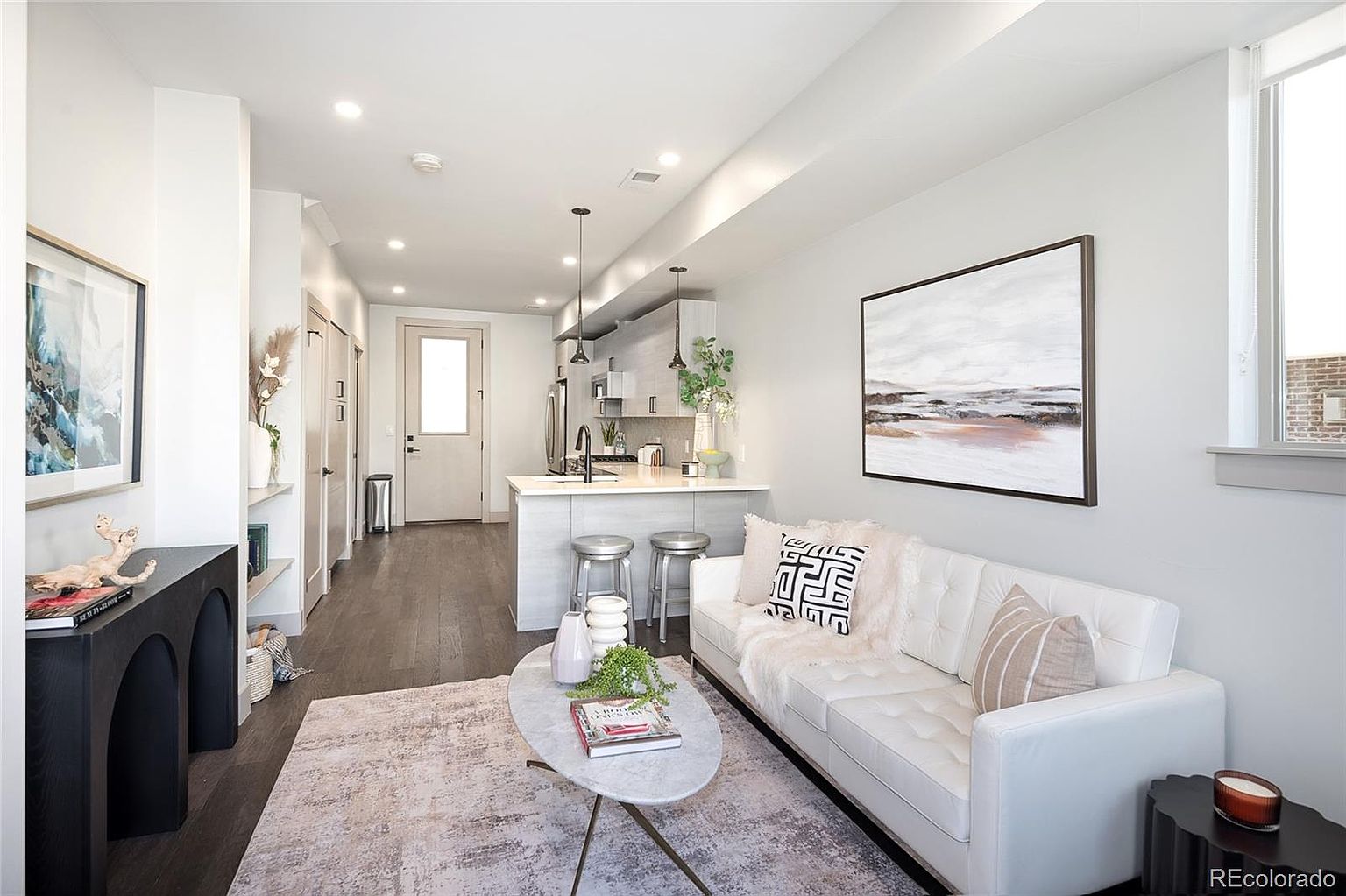 This is an interior shot of a modern living room that flows into a kitchen area. The living room features a white sofa with decorative pillows, a marble coffee table, and a black console table against the wall. The kitchen has light gray cabinets and a breakfast bar with stools, creating an open and inviting space.