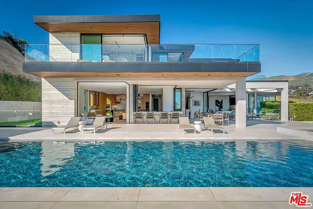This stunning contemporary luxury residence features a seamless indoor-outdoor flow with expansive glass walls that open onto a spacious stone patio. The property boasts a sparkling infinity-edge pool, multiple lounge areas, and a sleek, modern architectural design with wood accents and clean lines. The perspective captures the entire rear facade, highlighting the sophisticated integration of the home with its outdoor living space.