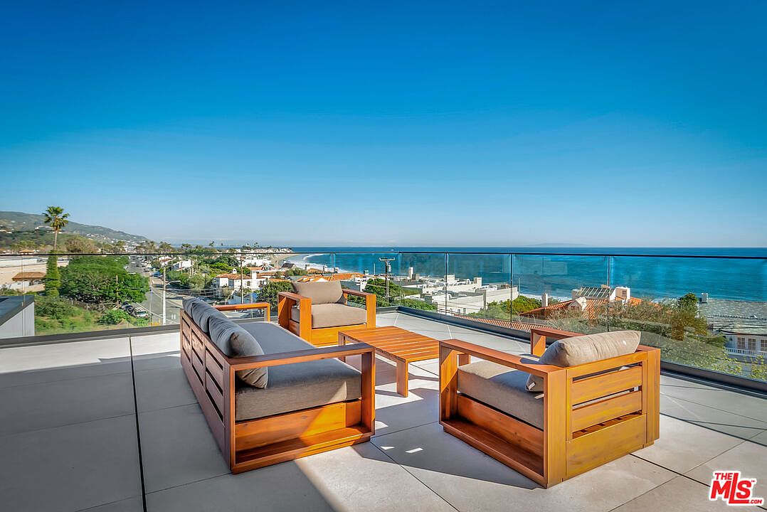 This stunning rooftop deck features modern wooden outdoor furniture, including a sofa and armchairs, arranged to take full advantage of the panoramic ocean views. The space is enclosed by a sleek glass railing, creating an unobstructed connection between the terrace and the coastal landscape below. The bright, sunny atmosphere and clean lines emphasize a luxurious, contemporary lifestyle perfect for entertaining.