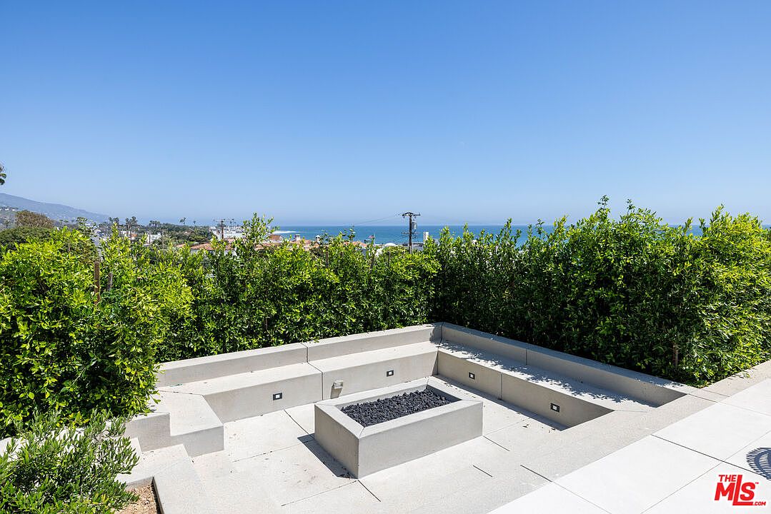 This outdoor space features a modern, built-in concrete fire pit surrounded by integrated bench seating, creating a sleek and minimalist lounge area. The patio is framed by lush, tall green hedges that provide privacy while framing a stunning, unobstructed panoramic view of the ocean and coastline. The perspective is eye-level, capturing the serene atmosphere of a high-end coastal retreat perfect for sunset gatherings.