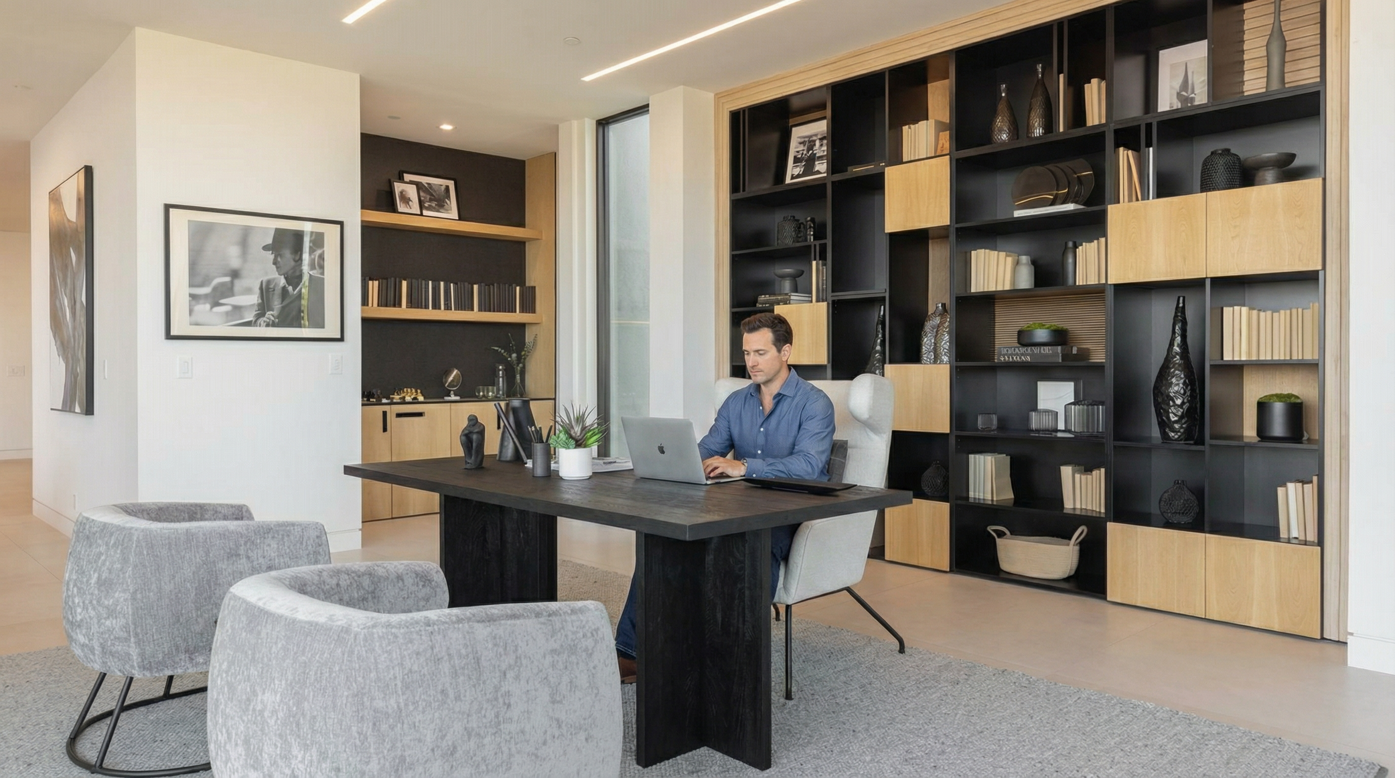 This sophisticated home office features a sleek, dark wood desk centered in the room, complemented by two modern grey upholstered armchairs. A large, floor-to-ceiling shelving unit with a mix of open black metal frames and light wood cabinetry provides ample storage and display space. The room is illuminated by minimalist recessed linear lighting, creating a clean, professional, and contemporary atmosphere.