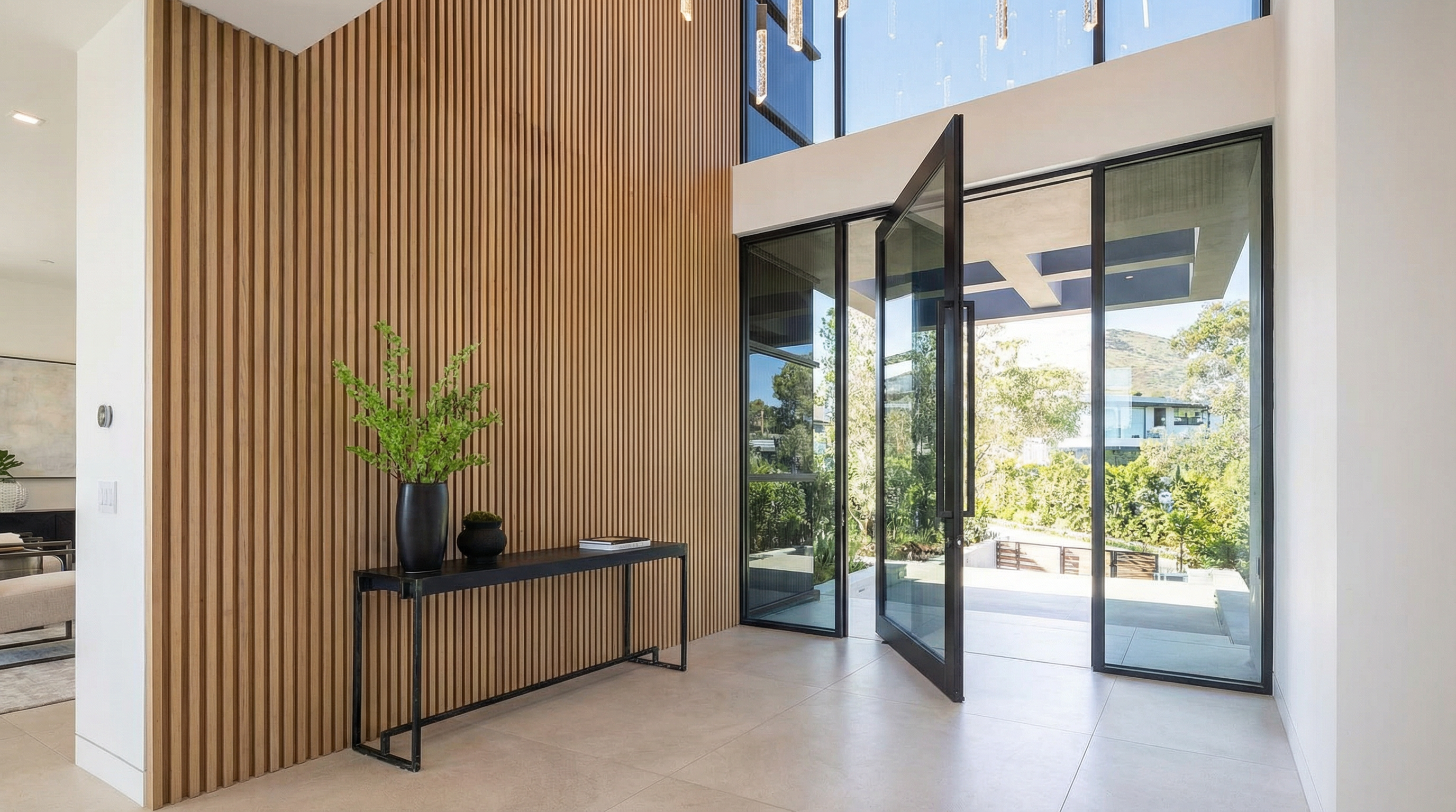 This grand, modern entryway features a dramatic floor-to-ceiling glass pivot door that floods the space with natural light. The interior is characterized by a striking wood-slat accent wall, a minimalist console table with a decorative plant, and a sophisticated, multi-tiered crystal chandelier hanging from the high ceiling. The clean lines, neutral tile flooring, and seamless transition to the outdoor landscape create an atmosphere of luxury and architectural elegance.