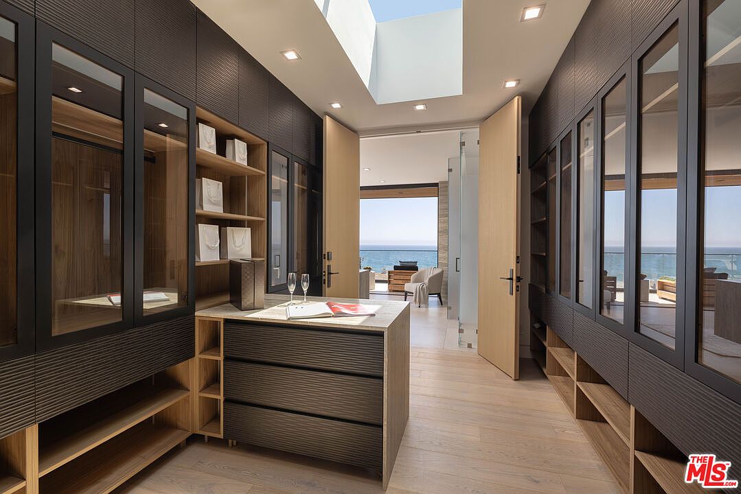 This luxurious walk-in closet features custom dark-textured cabinetry with glass-fronted doors and warm wood shelving, creating a sophisticated and organized aesthetic. A central island with a stone countertop provides a functional workspace, while a skylight above floods the space with natural light. The open doorway leads directly into a bright, airy living area with a stunning ocean view, emphasizing the seamless transition between indoor storage and coastal living.