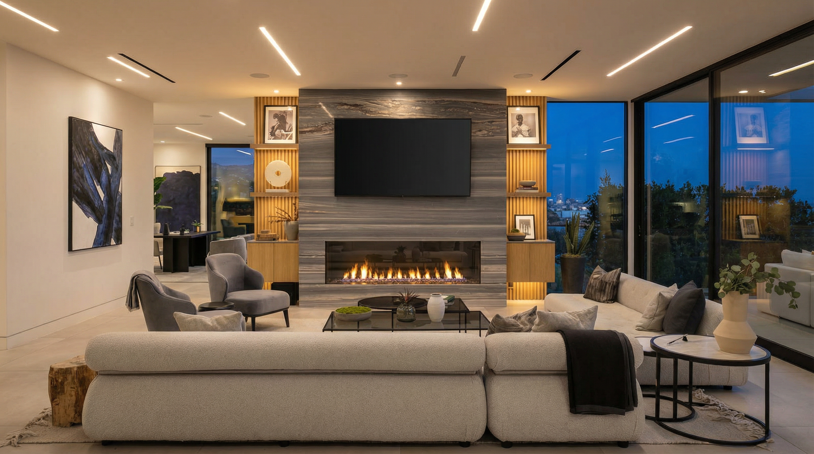 This sophisticated, open-concept living room features a striking floor-to-ceiling stone fireplace as its centerpiece, flanked by custom wood shelving. A large, plush sectional sofa and modern armchairs create a comfortable seating area, while expansive floor-to-ceiling glass walls offer a seamless transition to the outdoor scenery. The space is defined by its clean, contemporary aesthetic, recessed linear lighting, and a neutral color palette that emphasizes luxury and relaxation.