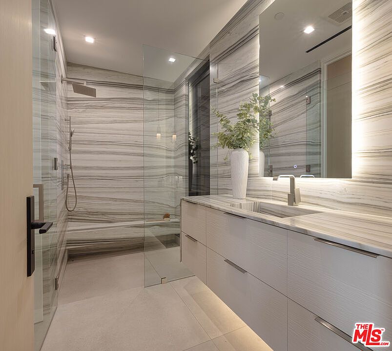 This luxurious primary bathroom features striking, book-matched marble walls with horizontal veining that create a sophisticated, high-end aesthetic. The space includes a floating vanity with integrated lighting, a sleek glass-enclosed walk-in shower with a built-in bench, and a modern, minimalist design. The perspective captures the seamless flow between the vanity area and the shower, emphasizing the clean lines and premium materials.