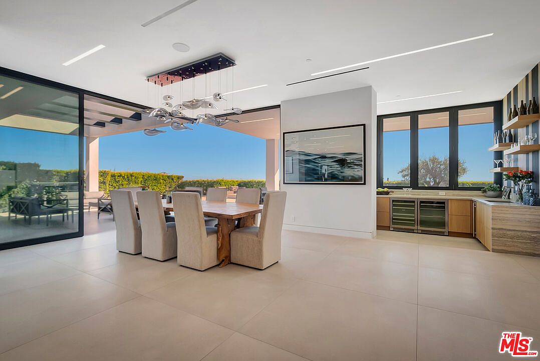 This sophisticated dining area features a natural wood table surrounded by six upholstered chairs, positioned beneath a striking, modern multi-pendant light fixture. The open-concept space seamlessly transitions to an outdoor patio through large sliding glass doors, while an adjacent wet bar area with wine refrigeration adds functionality and luxury. The room is characterized by its clean lines, neutral color palette, and expansive views, creating an airy and inviting atmosphere perfect for entertaining.