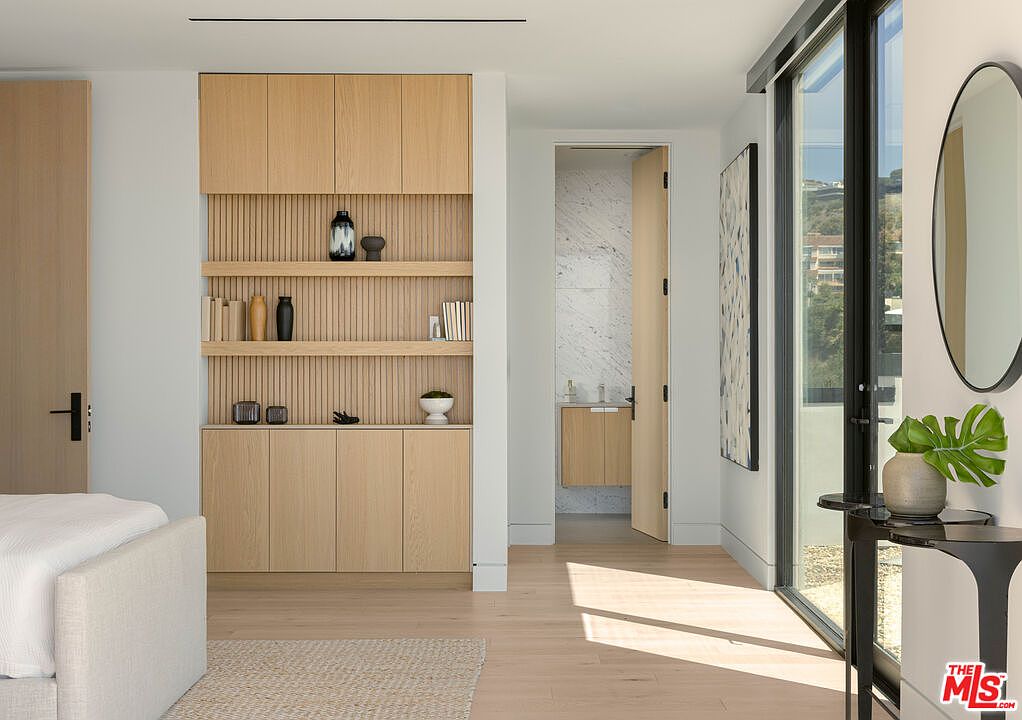 This modern primary bedroom features a sleek, light wood built-in shelving unit with integrated storage and vertical slat detailing. The room flows seamlessly into an en-suite bathroom with marble accents, while a large glass door provides abundant natural light and a view of the outdoors. The minimalist aesthetic is complemented by a neutral color palette and a contemporary circular mirror.